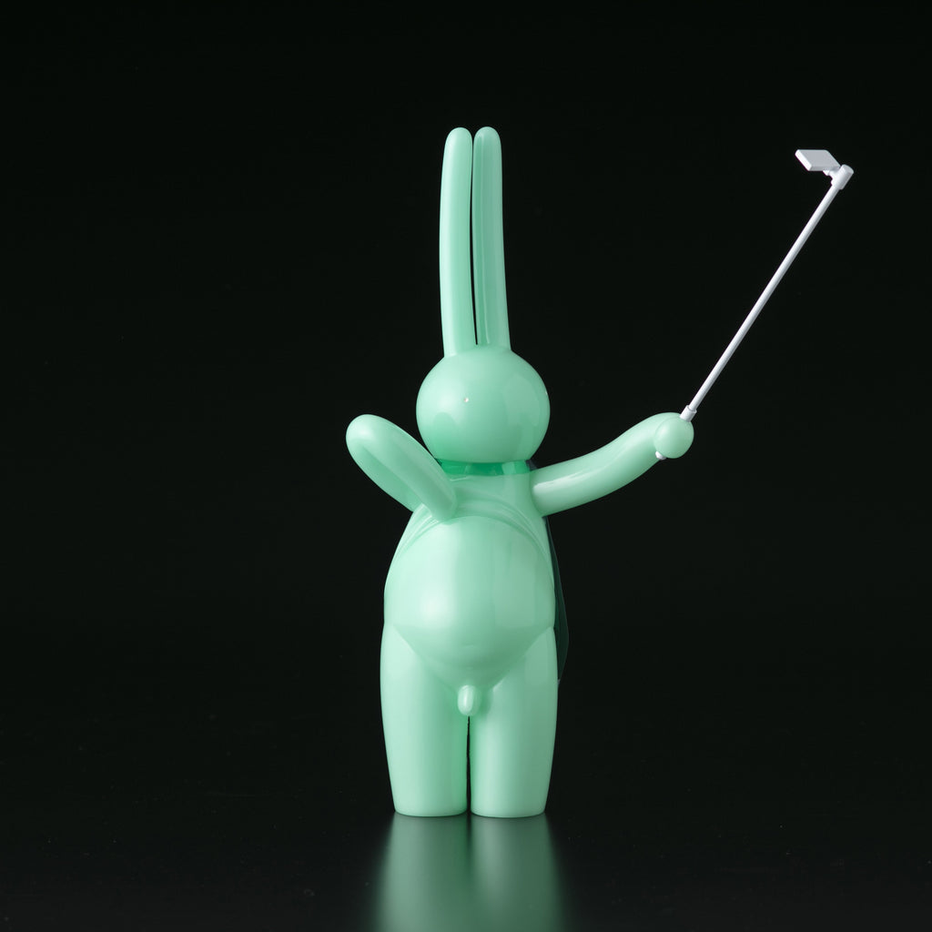 The Daily Flasher by mr. clement for Tomenosuke Shoten is a mint green, glow-in-the-dark vinyl rabbit with a selfie stick, facing away—a playful, abstract collectible that stands out against a black background.