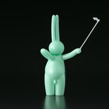 The Daily Flasher by mr. clement for Tomenosuke Shoten is a mint green, glow-in-the-dark vinyl rabbit with a selfie stick, facing away—a playful, abstract collectible that stands out against a black background.