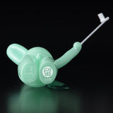 The Daily Flasher by mr. clement for Tomenosuke Shoten is a mint green plastic piece with a round base, long handle, and thin white rod, resembling a glow-in-the-dark vinyl toy, set against a black background.