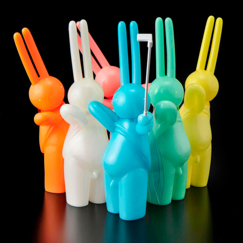 Six vibrant, bunny-shaped The Daily Flasher figures by mr. clement for Tomenosuke Shoten—including a glow-in-the-dark vinyl—stand together on a black background, with the blue one in front holding a selfie stick.