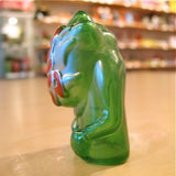 The Pop Soda Popsoda Finger Puppet - Clear Green stands on a light surface with blurred shelves behind it, recalling figures from a kaiju puppet show.