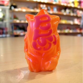 The Popsoda Finger Puppet - Orange by Pop Soda is a plastic figure in orange and pink with stylized text on its back, shown on a tabletop with blurred shelves of toys in the background.