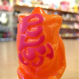 The Popsoda Finger Puppet - Orange by Pop Soda, featuring pink abstract markings, sits on a table as shelves and objects blur softly in the background.