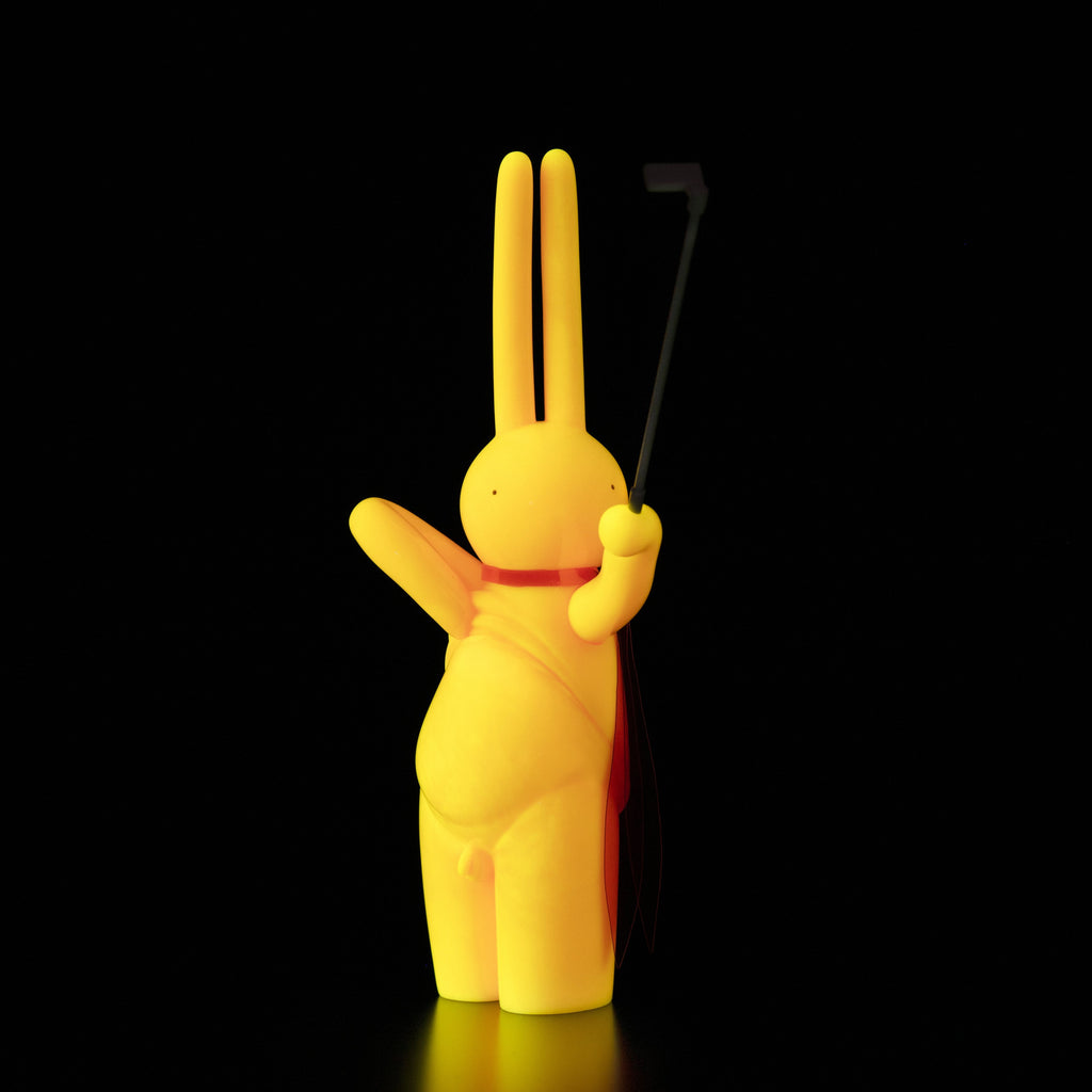 The Daily Flasher by mr. clement, from Tomenosuke Shoten, is a yellow sofubi rabbit figurine with a red cape holding a selfie stick against a black background, reflecting the toy brand’s playful spirit.