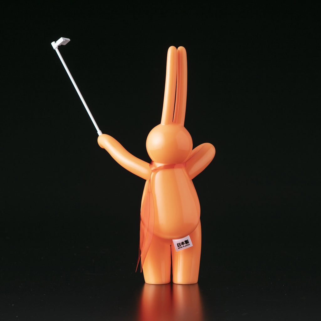 The Daily Flasher by mr. clement from Tomenosuke Shoten is a glow-in-the-dark orange rabbit figurine with a selfie stick, set against a black background—a playful vinyl collectible for any collection.