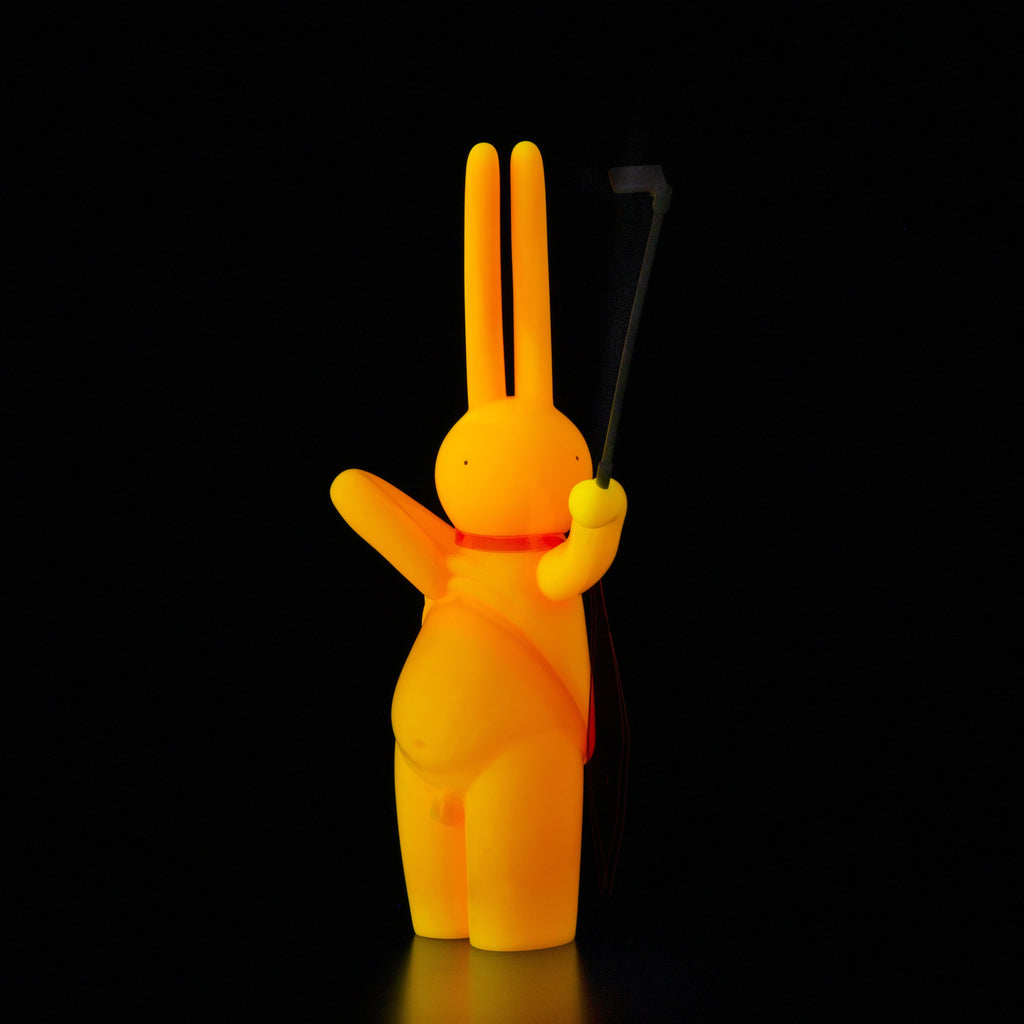 The Daily Flasher by mr. clement, from Tomenosuke Shoten, is a yellow sofubi bunny figurine with a round belly and red strap, holding a selfie stick against a black background.
