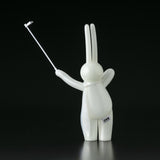 Against a black background, The Daily Flasher by mr. clement—an exclusive glow-in-the-dark vinyl rabbit from Tomenosuke Shoten—poses with a selfie stick, its side adorned with Japanese text.