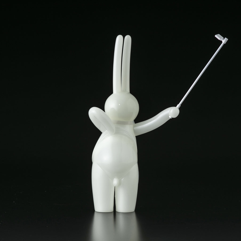 The Daily Flasher by mr. clement for Tomenosuke Shoten is a glossy white bunny figurine, standing upright with a raised right arm holding a selfie stick against a black background.