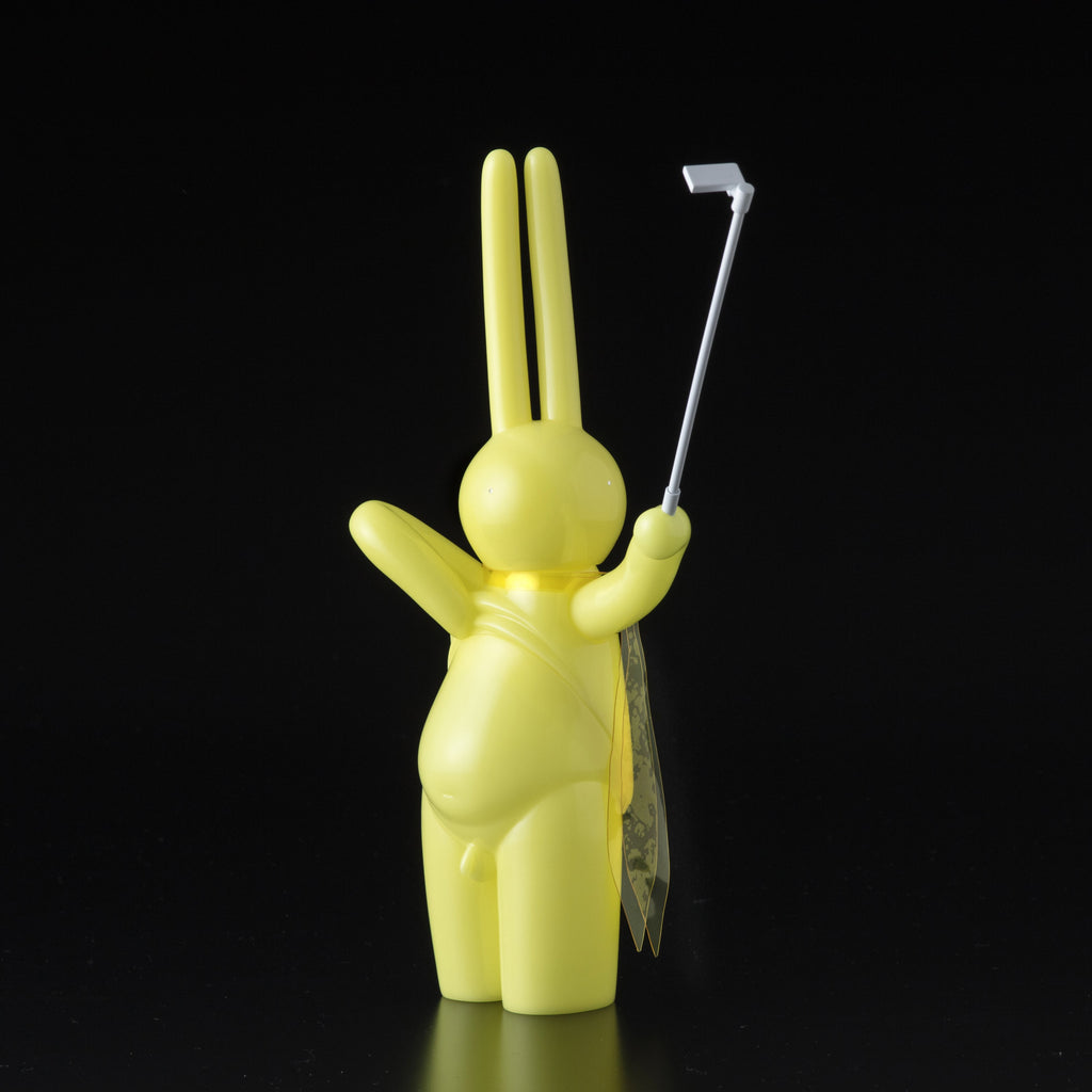 The Daily Flasher by mr. clement for Tomenosuke Shoten is a yellow, long-eared bunny vinyl figure with a selfie stick and translucent cape that glows in the dark—adding playful charm to any collection.