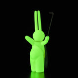 The Daily Flasher by mr. clement is a glowing green bunny vinyl toy with a simple face, holding a stick upright against a black background, available at Tomenosuke Shoten.