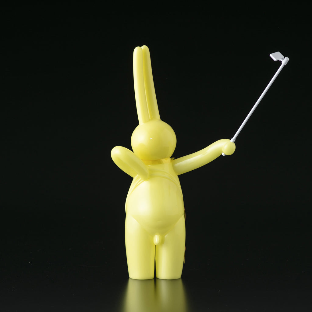 The Daily Flasher by mr. clement, a glow-in-the-dark yellow vinyl rabbit from Tomenosuke Shoten, is shown with a selfie stick in its right hand against a plain black background.