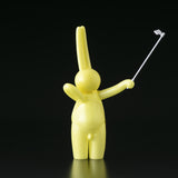 The Daily Flasher by mr. clement, a glow-in-the-dark yellow vinyl rabbit from Tomenosuke Shoten, is shown with a selfie stick in its right hand against a plain black background.