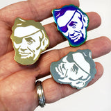 A hand displays three Rotofugi Pirate Abe Enamel Pins—Abraham Lincoln’s face with an eyepatch, in gold, silver, and iridescent finishes. Unique mascot pins from top designer toy brand Rotofugi.